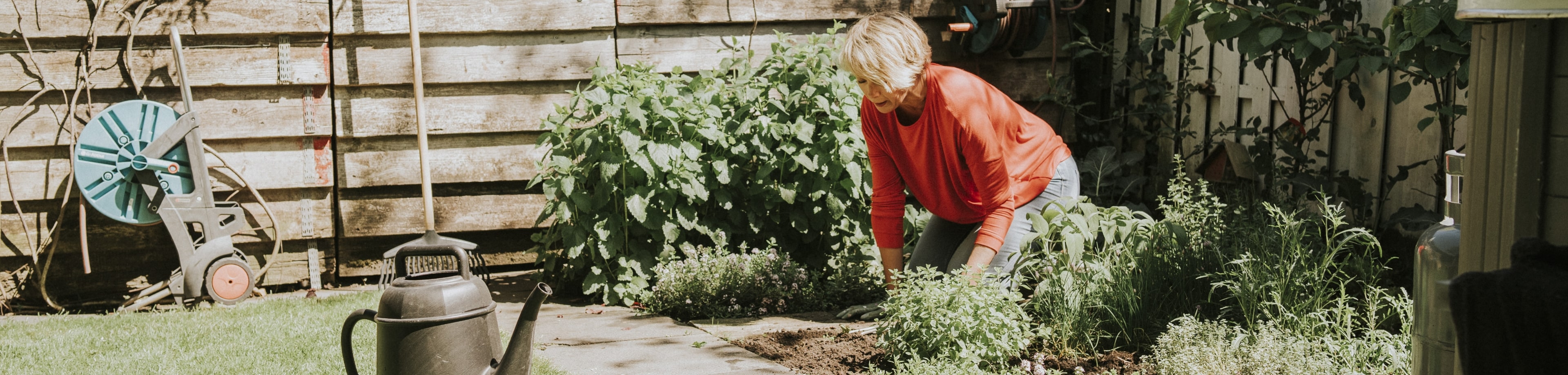 Een vrouw met blond haar werkt in de tuin. Ze zit op haar knieën en harkt de tuingrond los. Naast haar staan diverse soorten tuingereedschap.