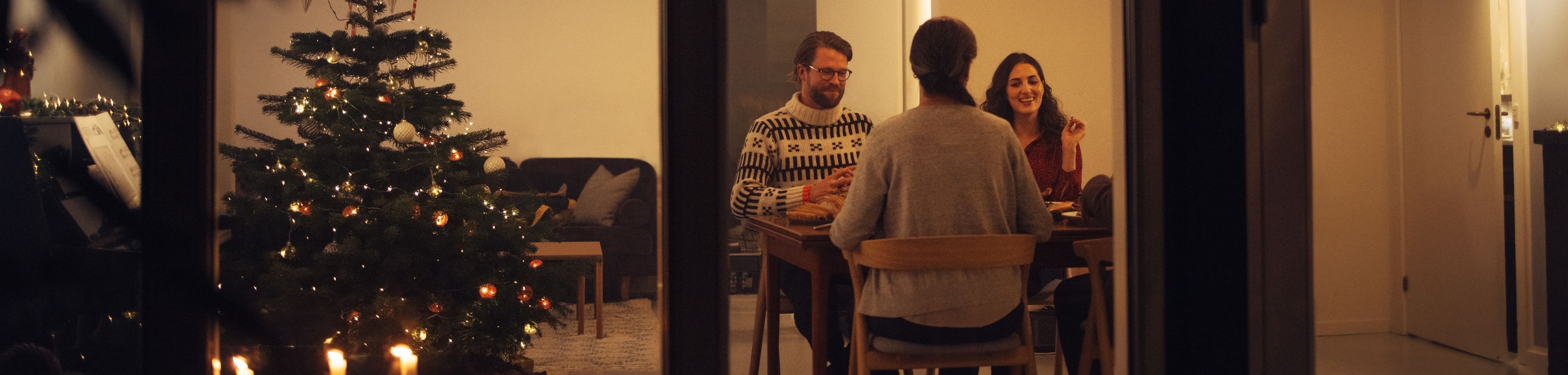 4 mensen zitten binnenshuis aan een keukentafel, naast een rijkversierde kerstboom.