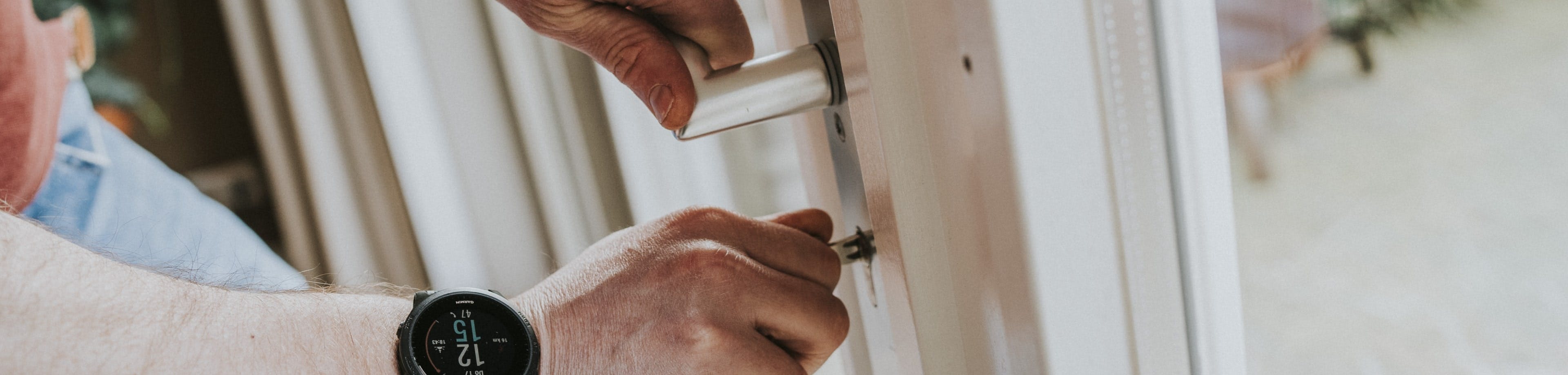 De handen van een persoon; de linkerhand pakt een deurklink vast, de rechterhand steekt een sleutel in het slot daar direct onder.