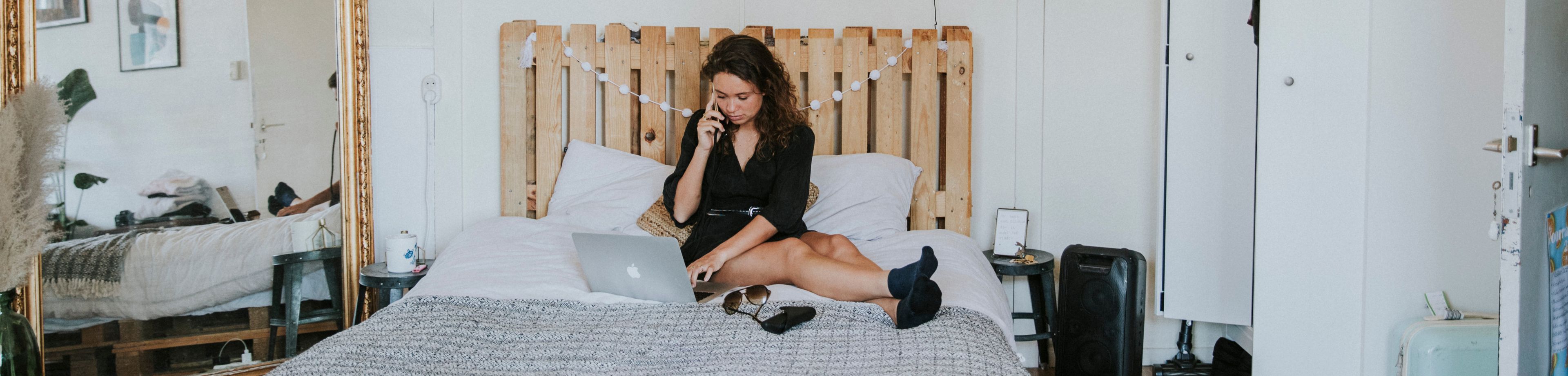 Jonge vrouw zit op het bed in haar studentenkamer en belt terwijl ze op de laptop bezig is.