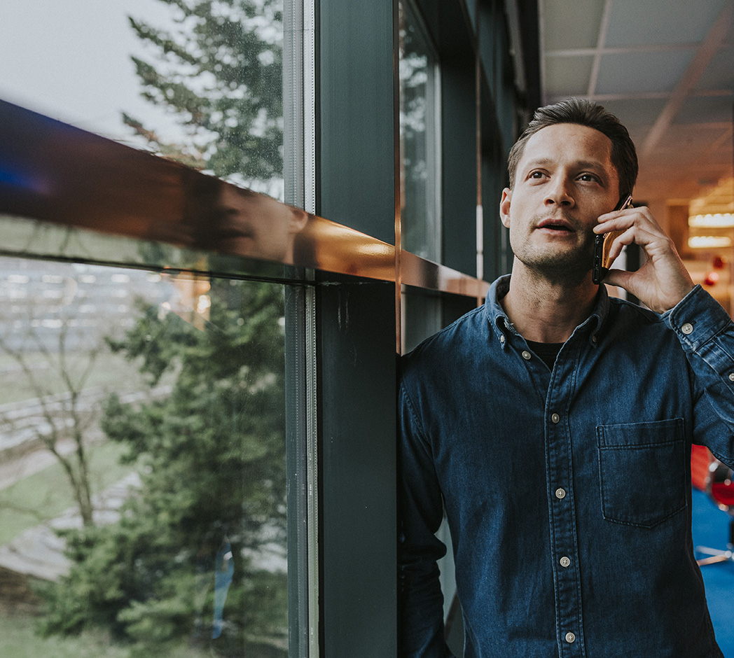 Een persoon staat met een schouder tegen een raamkozijn, en is in gesprek via een mobiele telefoon.