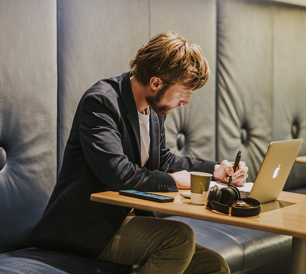Een persoon zit aan een houten tafeltje te werken. Op tafel staat een laptop en er liggen een schrijfblok en een koptelefoon. De persoon schrijft iets op in het schrijfblok.