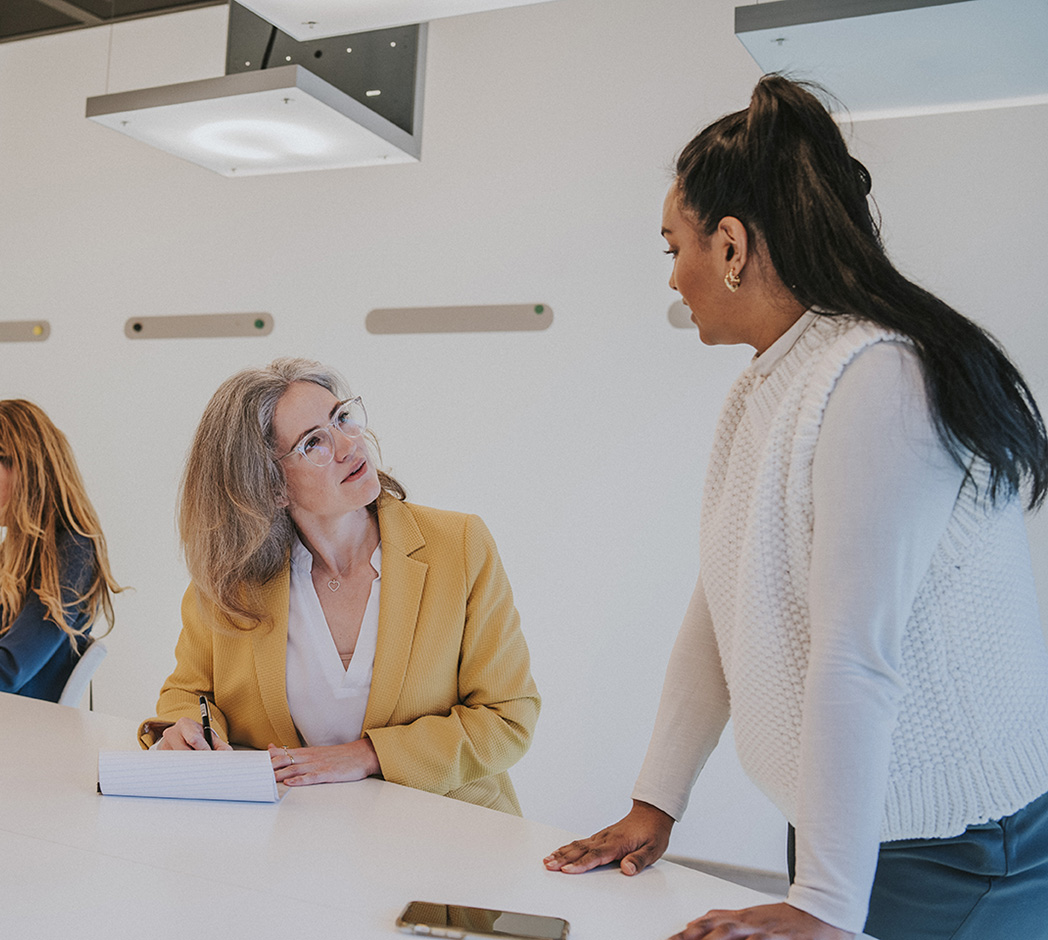 2 mensen zijn met elkaar in gesprek in een ruimte met een witte wand. 1 persoon zit, de ander staat. Ze kijken elkaar aan terwijl ze aan het praten zijn.