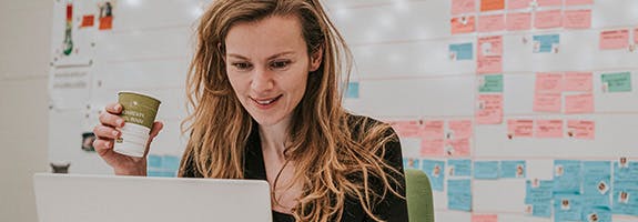 Een vrouw met lang, loshangend blond haar zit achter een laptop. Ze kijkt op het scherm en houdt een kartonnen koffiebeker in haar hand. Op de achtergrond: veelgekleurde notitieblaadjes op een witte wand.